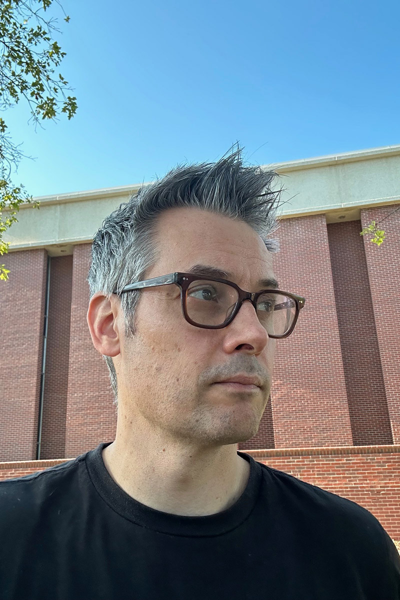 Headshot of Andrew Falkowski outdoors, wearing glasses and a black shirt, with a brick building and blue sky in the background.