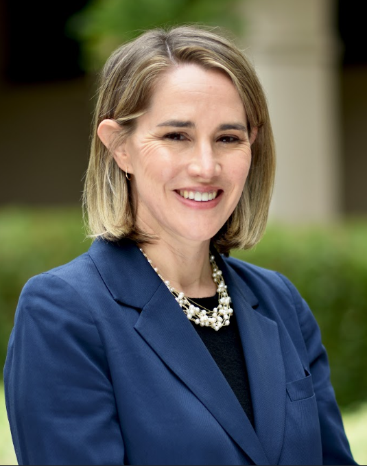 Person smiling, wearing a blue blazer and a decorative necklace, standing outdoors.
