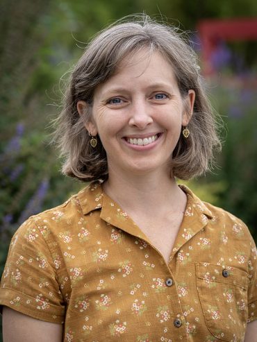 Portrait of a smiling individual with short hair, wearing a yellow floral shirt in front of a garden backdrop.