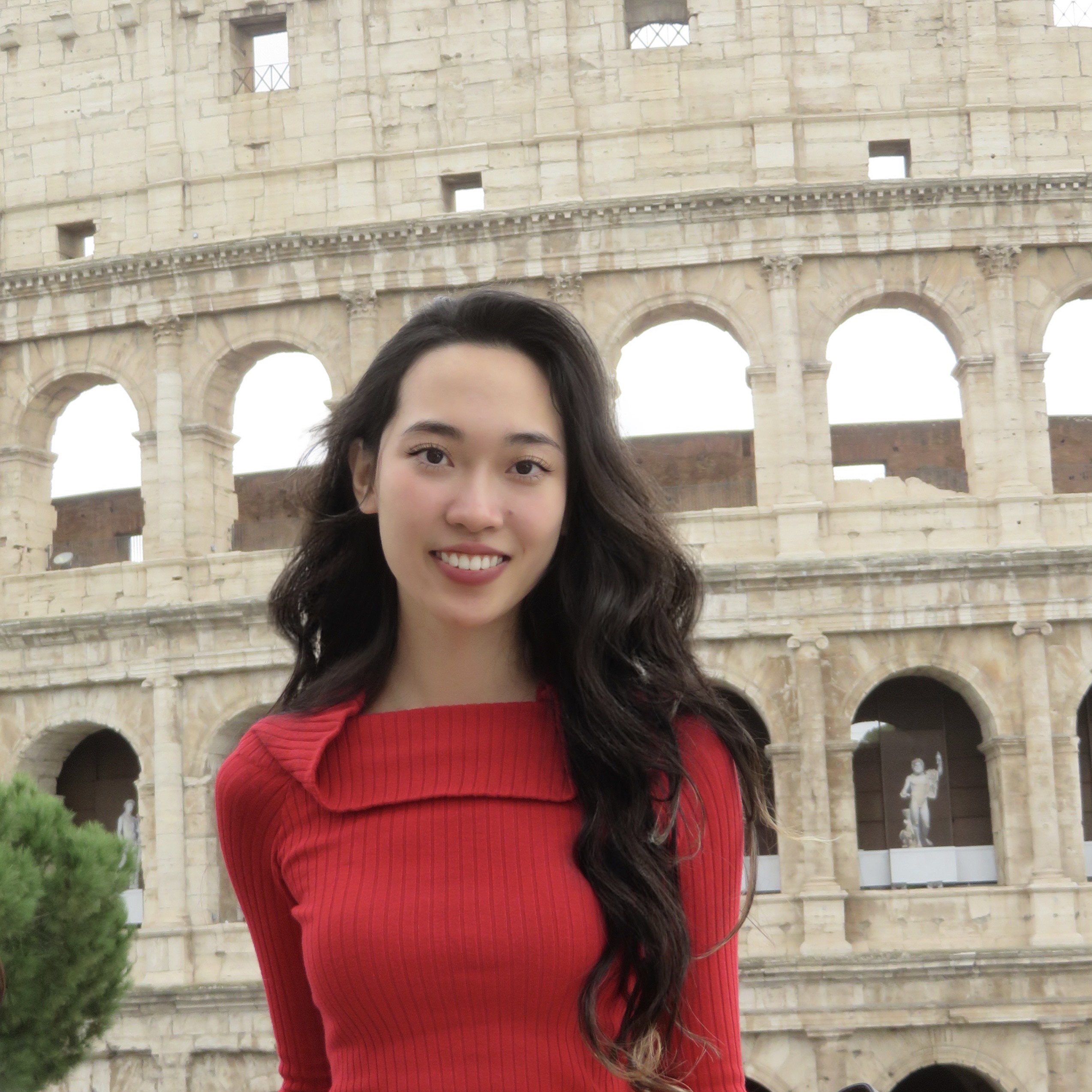 Rebecca Zhang A smiling woman in a red sweater stands in front of the Colosseum in Rome. The ancient structure's arches create a historic backdrop, conveying travel joy.