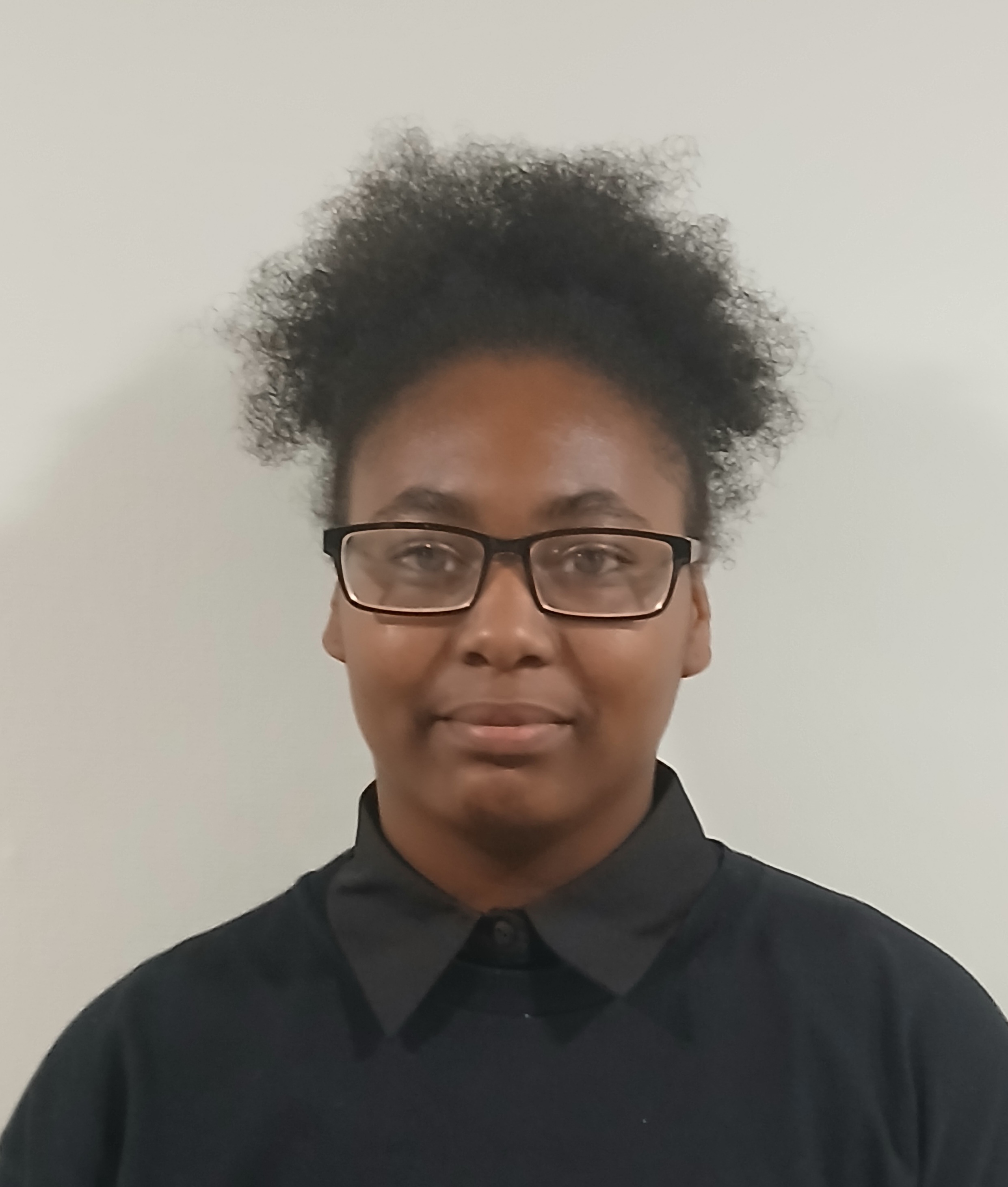 Ynuel Spencer A person with curly hair and glasses wears a black collared shirt. They stand against a plain white background, conveying a neutral expression.