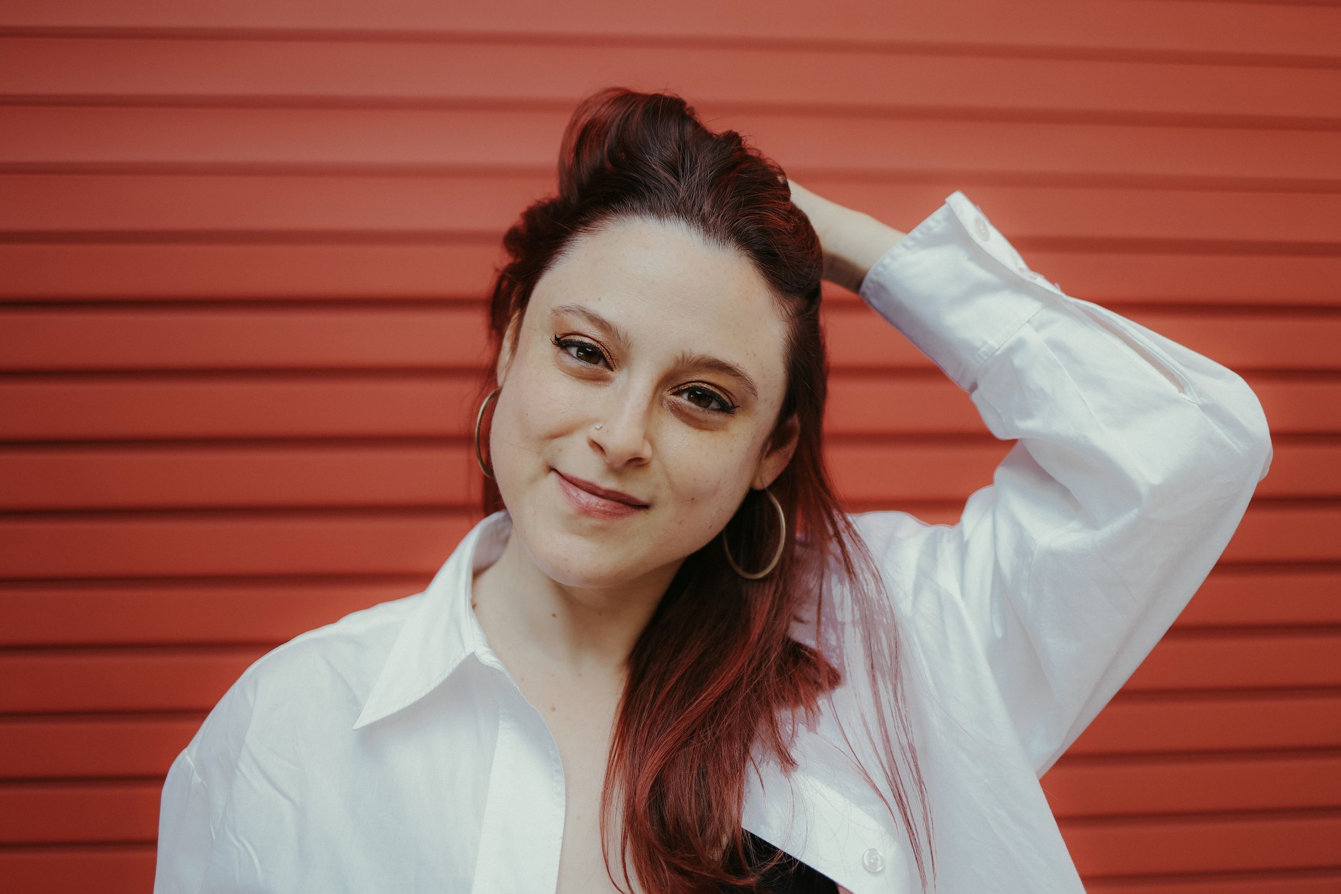 Anna Cahn Stony Brook University Art History Phd Candidate posing in front of a red background wearing hoop earrings and white button down shirt with her left hand in her hair.