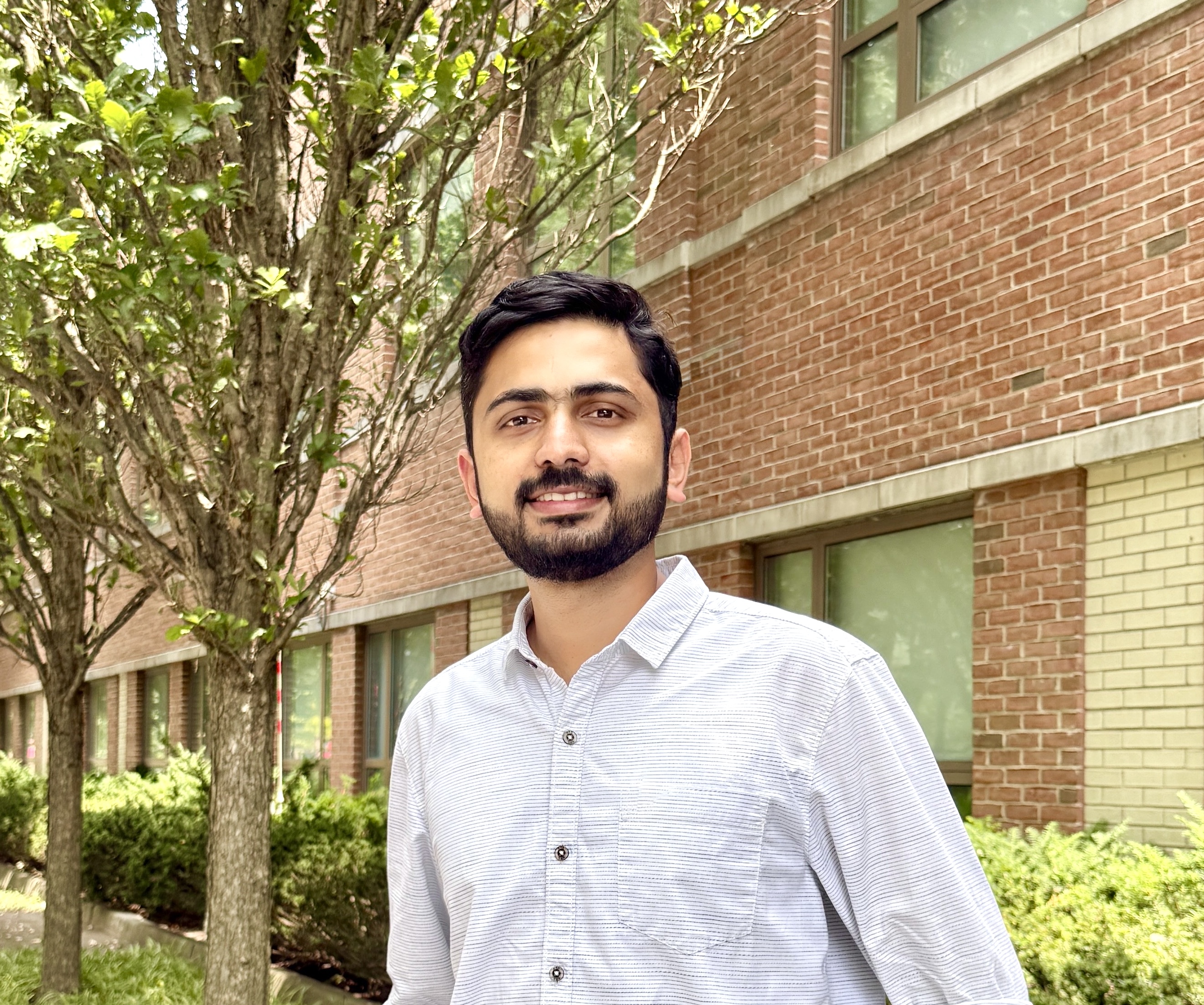 Donal Thomas  Person in a light blue shirt smiling and standing outdoors in front of brick buildings and green trees.