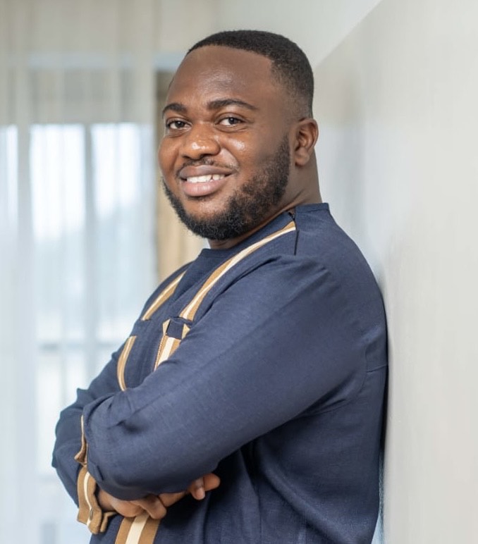 George Osei Portrait of a smiling person in traditional attire leaning against a wall.