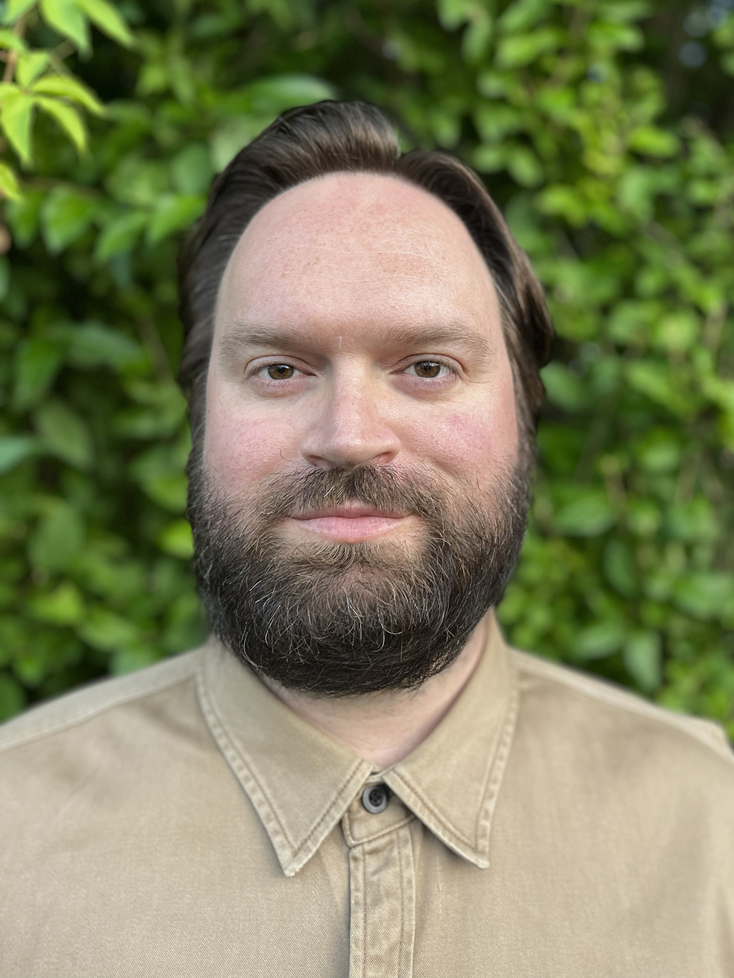 Portrait of an individual with shoulder-length brown hair and a beard, wearing a beige shirt, standing in front of a leafy green background.