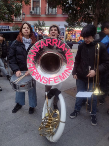 Three musicians playing in a street band, with a drum, a large horn, and a trombone. A banner on the horn reads "Fanfarria Transfeminista.