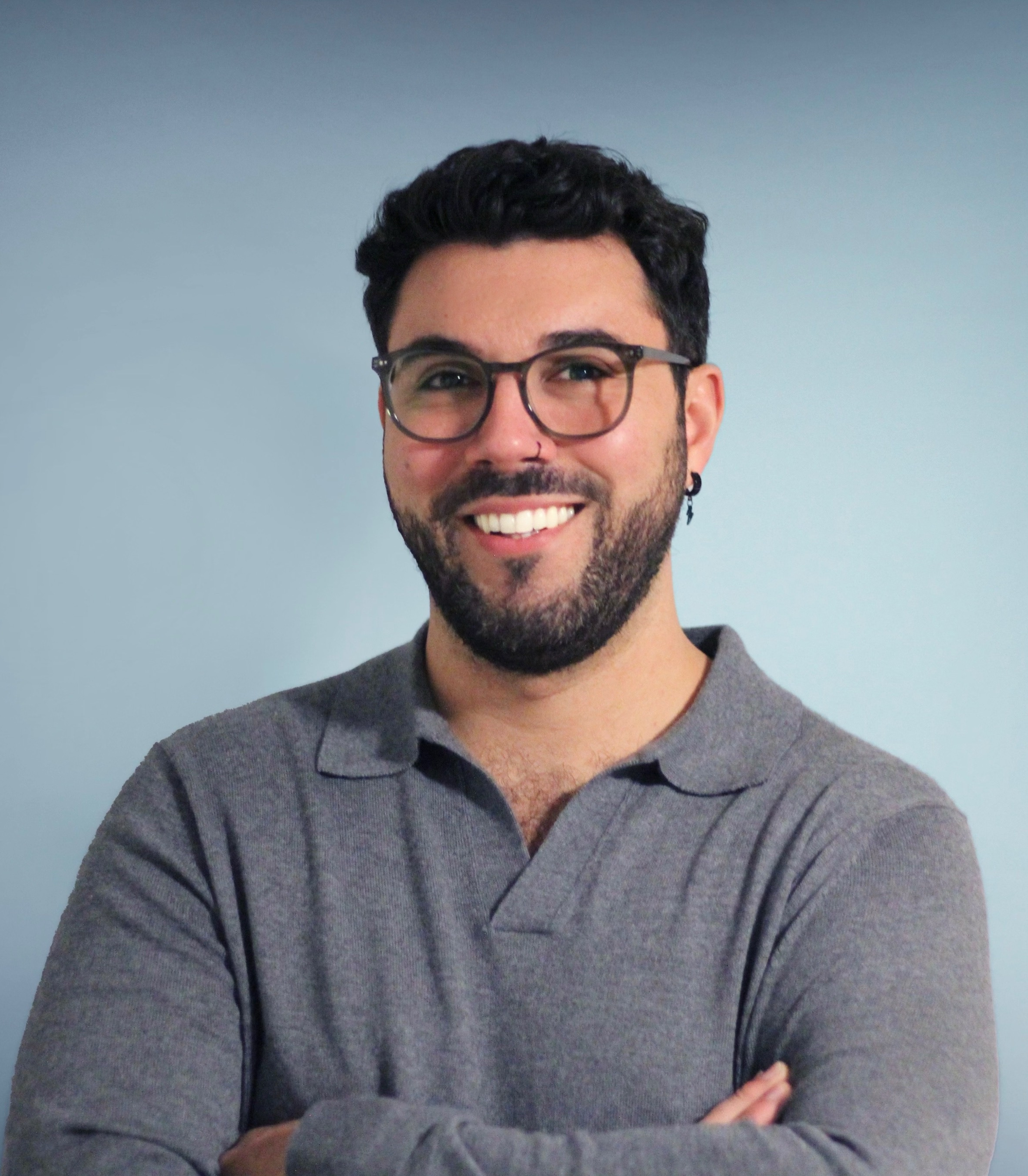 A smiling man with glasses and a beard stands against a light blue background. He is wearing a gray, long-sleeved, collared shirt and has his arms crossed."