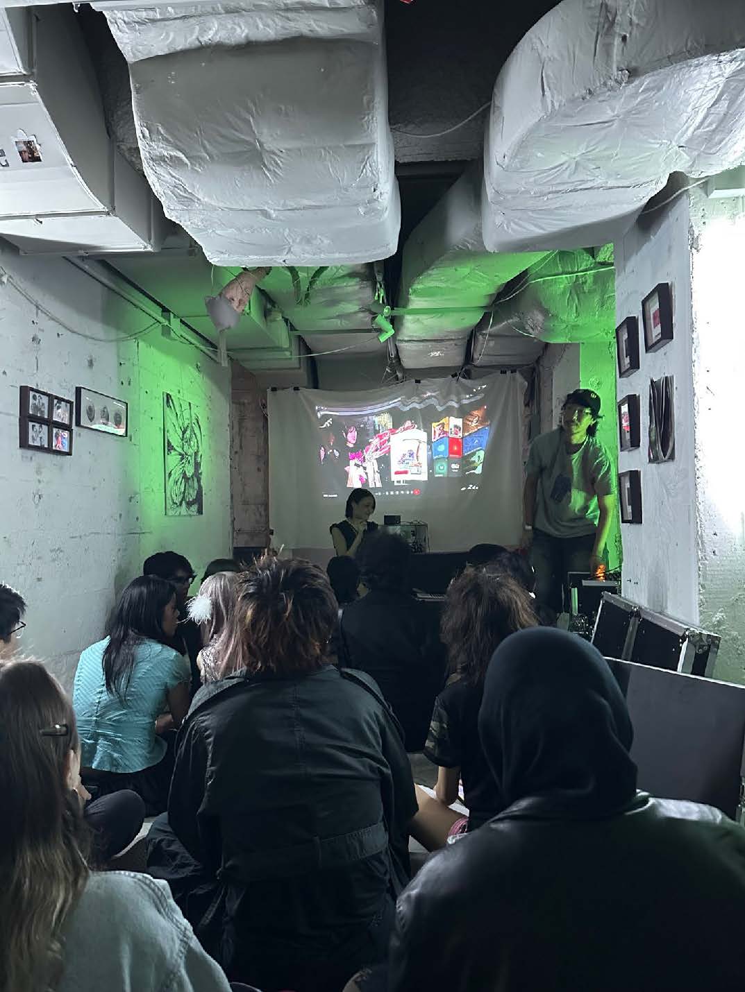 An audience seated on the floor in a small, densely packed room, watching a presentation projected on a screen. The room is dimly lit with art on the walls and a speaker standing near the projection.
