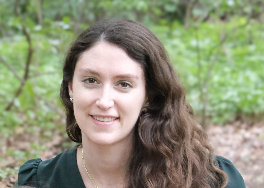 A woman with long, wavy brown hair smiles softly while sitting outdoors. The background features a lush, green forest, conveying a calm and serene atmosphere.