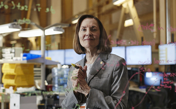 Esther Takeuchi Woman in a gray suit stands confidently in an office filled with computers, writing equations on a transparent board. The atmosphere is focused and analytical.