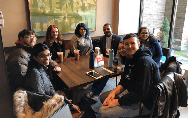 A group of eight people are gathered around a dark wooden square table in a brightly lit cafe or common area. They are smiling and looking toward the camera.  Seated on the left side: A young man in a tan puffer jacket and a young woman with long dark hair and glasses wearing a grey sweater.  Seated along the back: A woman with curly auburn hair in a black top, a woman with dark hair in a grey sweater, and a man in a blue blazer and white collared shirt.  Seated on the right side: A young woman in a blue puffer jacket, a woman partially obscured, and a young man in a black hooded sweatshirt and jeans in the foreground.  The table holds several iced coffee drinks, a white Starbucks cup, two reusable water bottles, a smartphone, and a small white sign with red text that reads "Blood Drive." A large abstract painting with green and yellow tones hangs on the wall behind them, and a large window to the right shows a brick building and a small evergreen tree outside.
