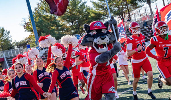 Woflie running onto football field with cheerleaders and players